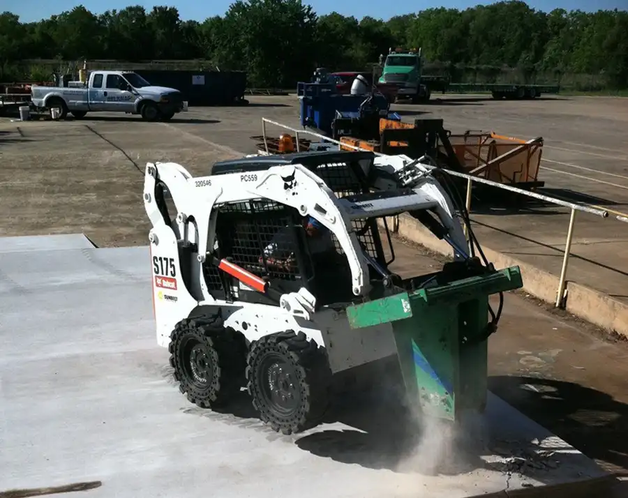 A Bobcat skid steer working a slab, a dumpster and pickup staged behind it