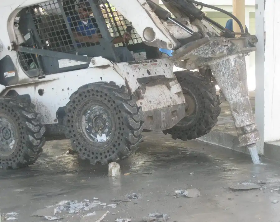 A skid steer running a hydraulic breaker attachment indoors with spoil piling up around it