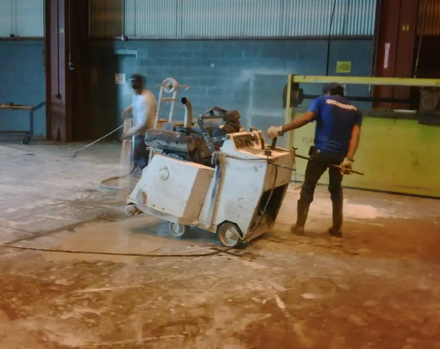 A walk-behind slab saw under water spray on a warehouse floor with two operators