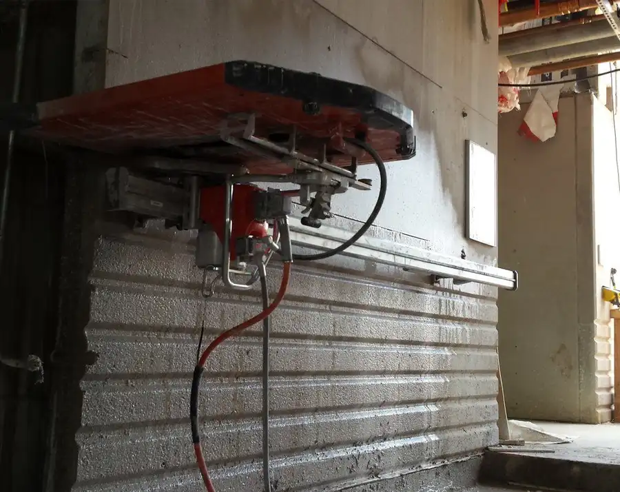 A track-mounted wall saw clamped high on a warehouse wall with hydraulic lines running to the head