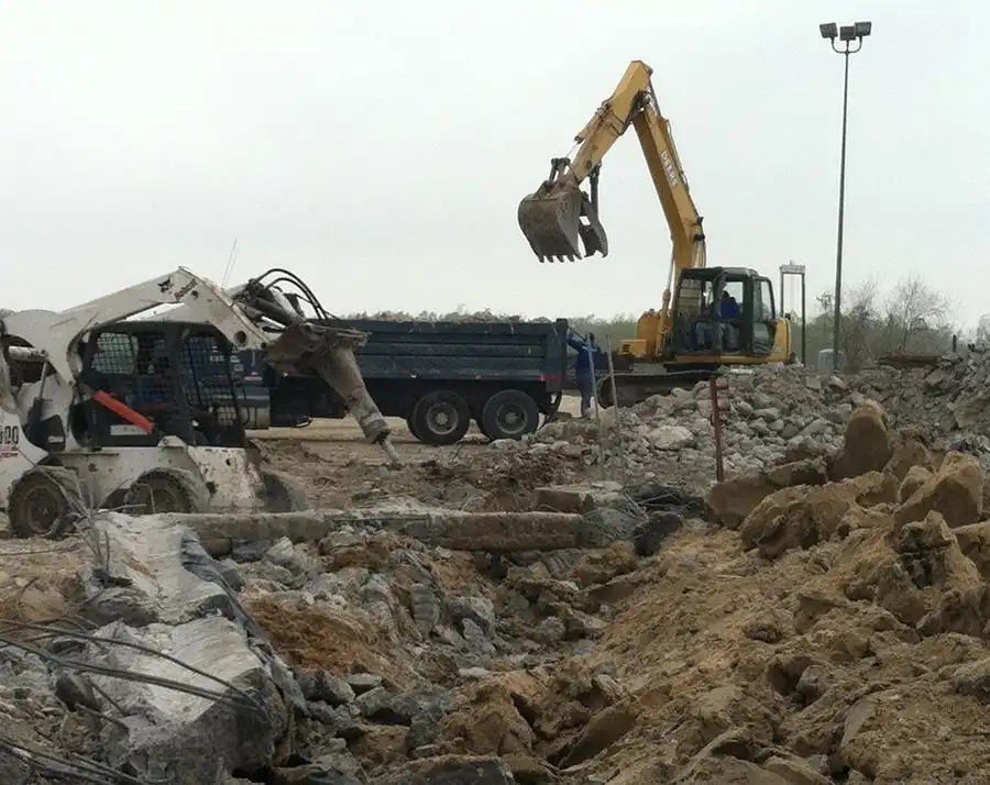 An excavator and a skid-steer breaker working a broken-concrete field, a dump truck staged to load out
